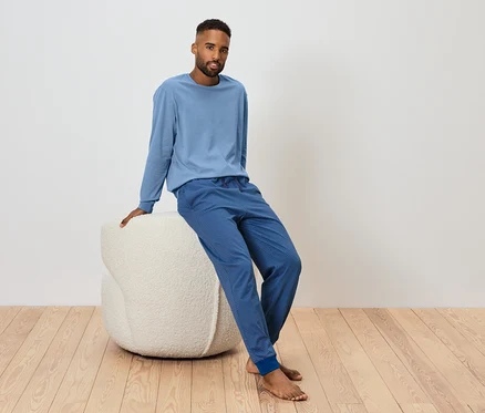 Un homme est assis sur un pouf blanc et porte un t-shirt bleu à manches longues et un pantalon de survêtement bleu.