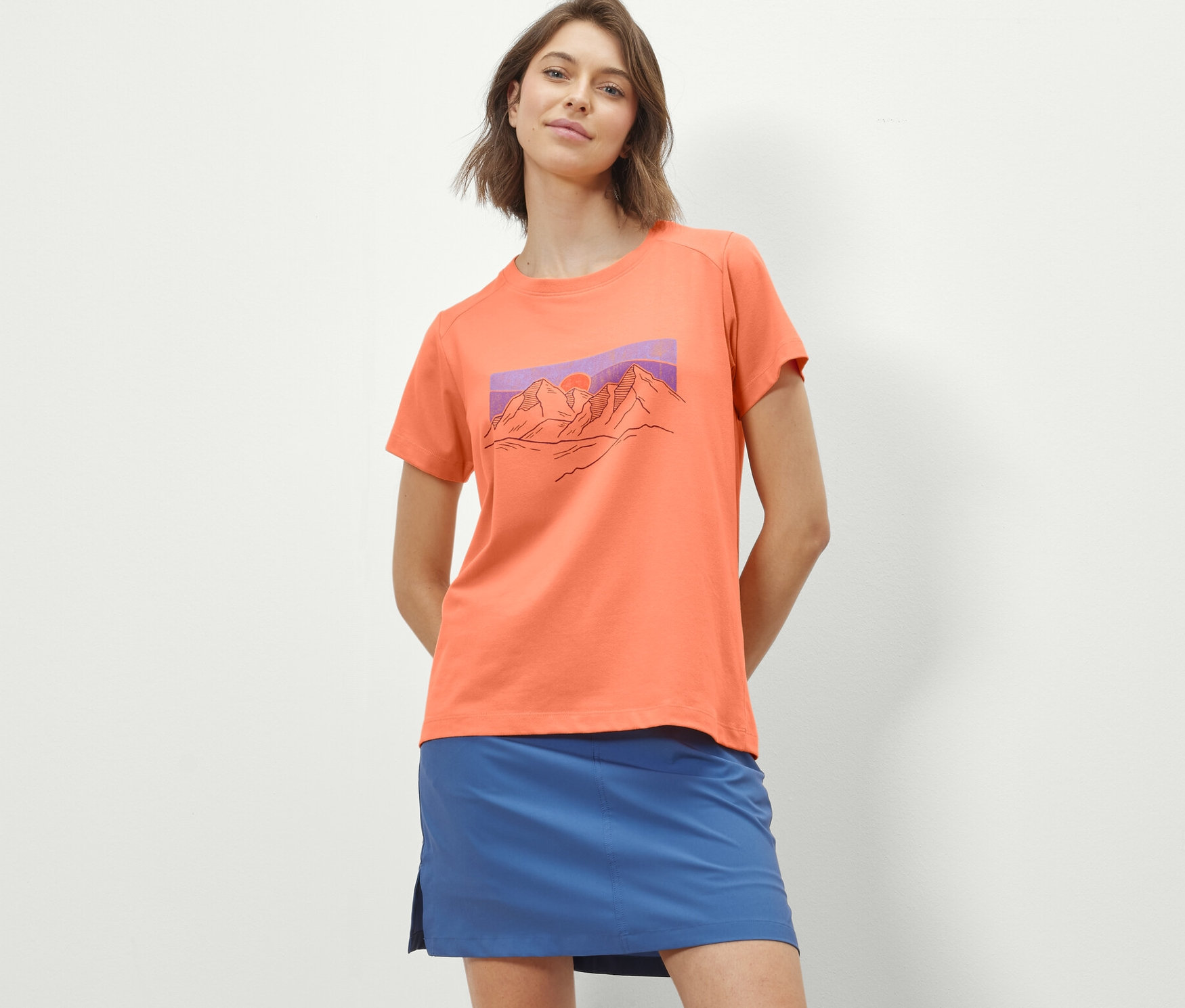 Une femme porte un t-shirt orange avec un imprimé et une jupe bleue.