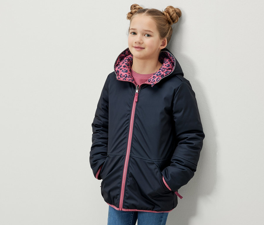 Une fille pose en veste softshell et matelassée 2-en-1 pour enfant avec capuche, fermeture éclair rose et bordures.