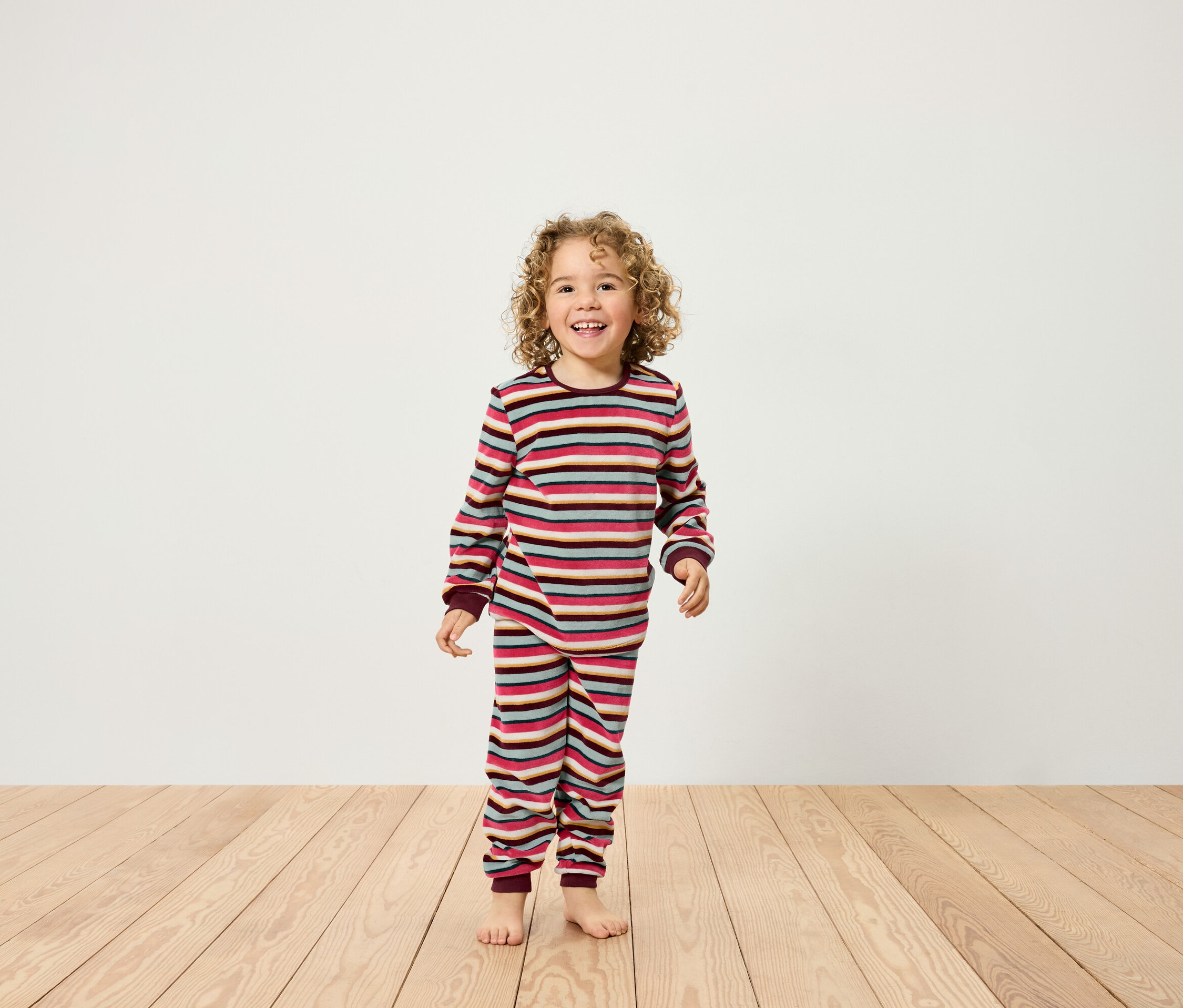 Un enfant souriant aux cheveux bouclés se tient sur un plancher en bois et porte un pyjama rayé.