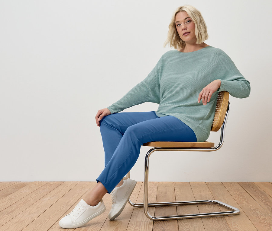 Une femme blonde est assise sur une chaise, portant un pantalon stretch bleu et un pull en maille fine sauge.