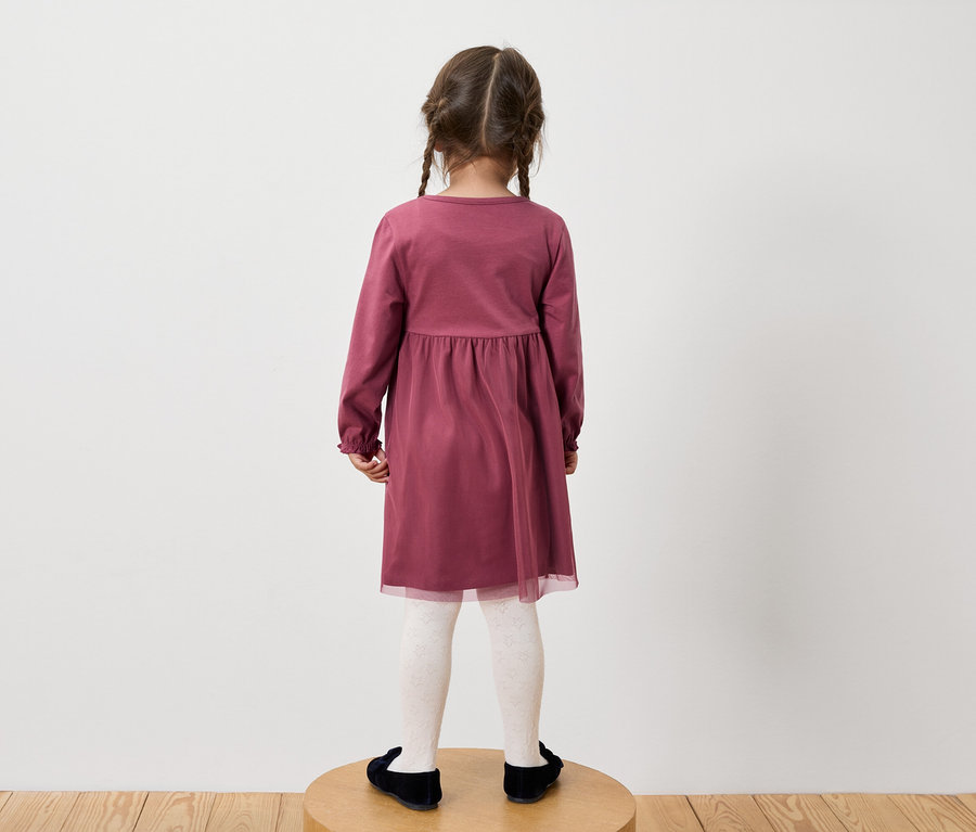 Une fillette portant une robe bordeaux à manches longues, des collants blancs à motifs et des chaussures noires, dos à l'appareil photo.