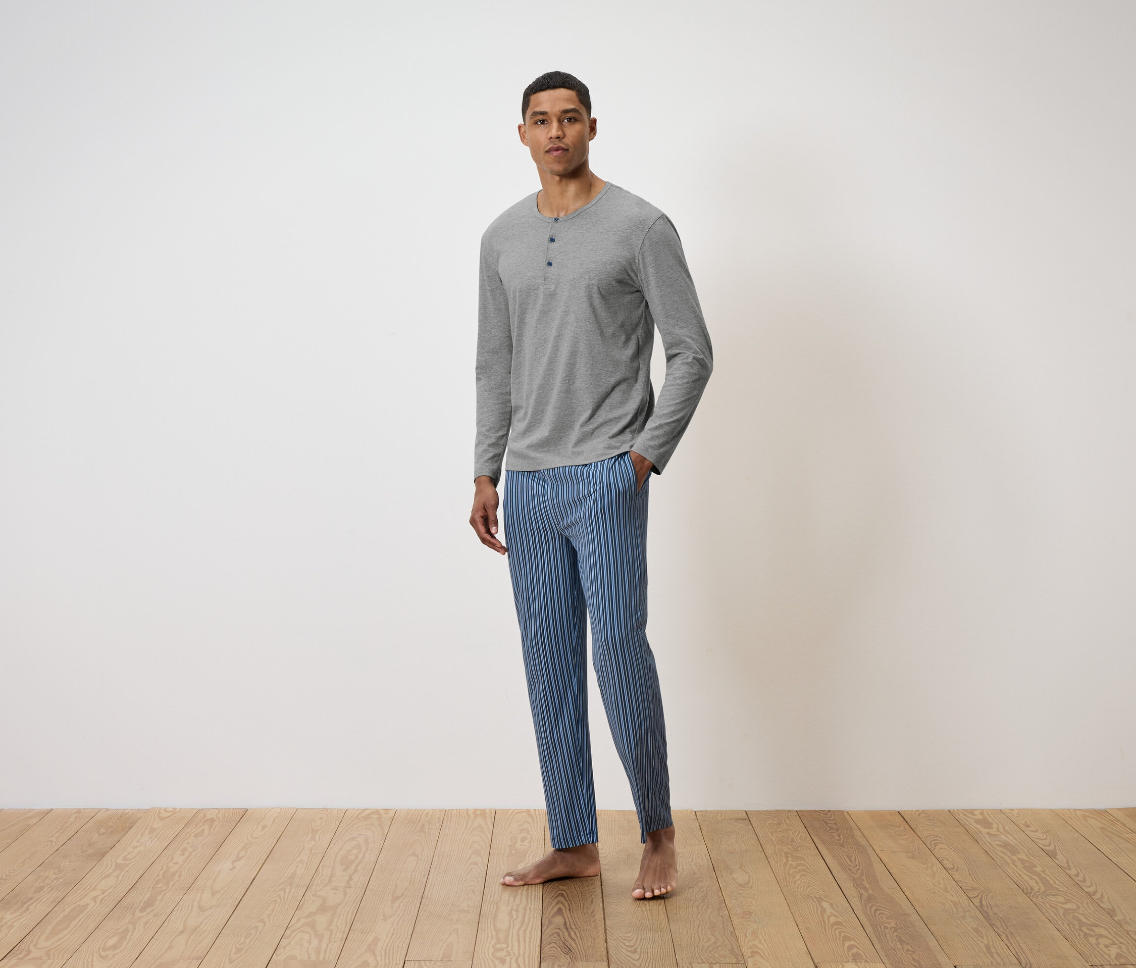 Un homme debout sur un plancher en bois, portant un t-shirt gris à manches longues et un pantalon rayé bleu.