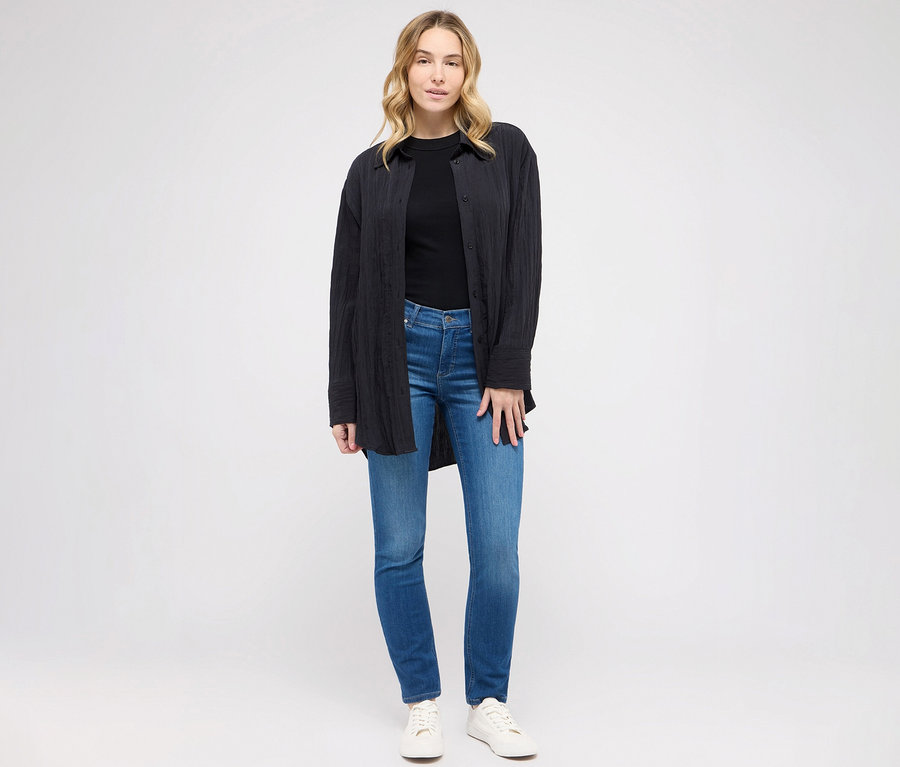 Femme posant en jean Angels « Cici » coupe étroite, bleu moyen, t-shirt noir et chemisier.