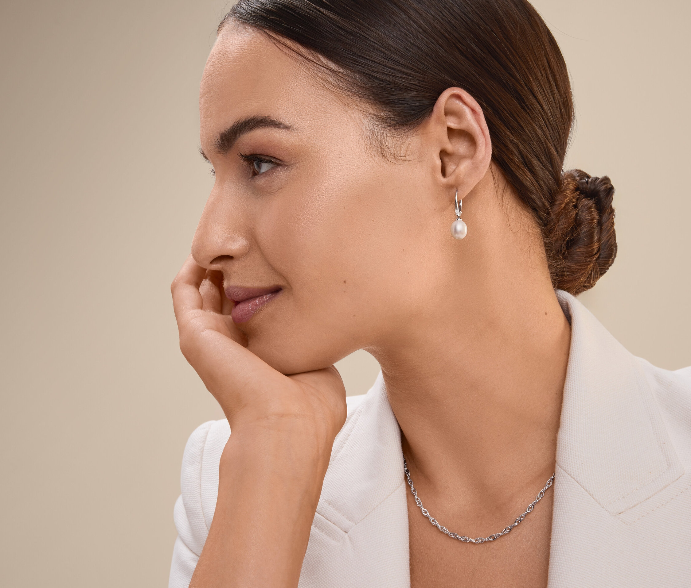 Femme aux cheveux bruns relevés en chignon, menton appuyé sur la main, portant une veste blanche, un collier et des boucles d'oreilles.