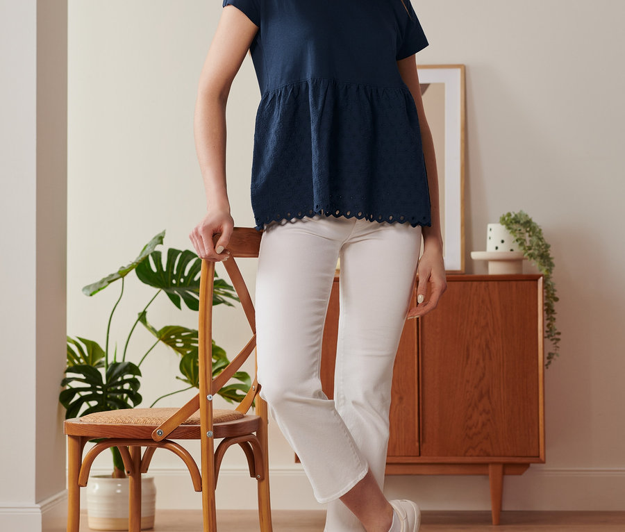 Une femme appuyée sur une chaise porte un t-shirt bimatière à broderie bleu foncé et un pantalon blanc.