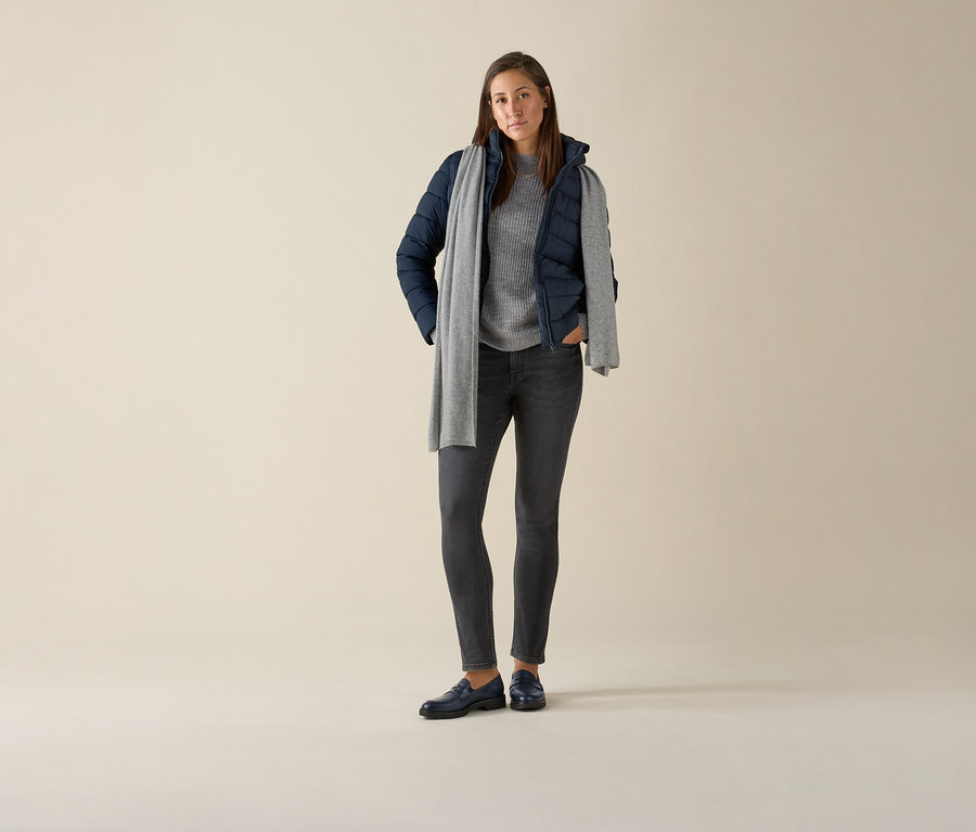 Une femme debout portant un jean slim – Coupe « Emma », denim gris, un pull gris, une veste bleu marine et une écharpe grise.
