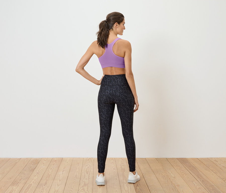 Une femme se tient dos à la caméra, portant un soutien-gorge de sport violet et un legging noir.