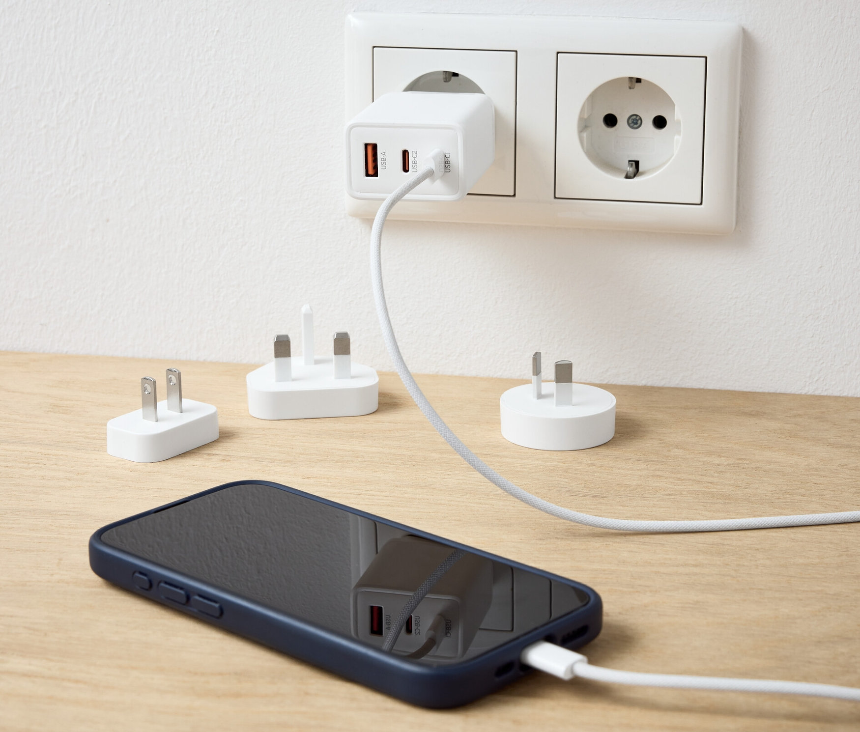 Un adaptateur de voyage USB blanc branché sur une prise de courant charge un téléphone portable. D'autres adaptateurs sont sur la table.