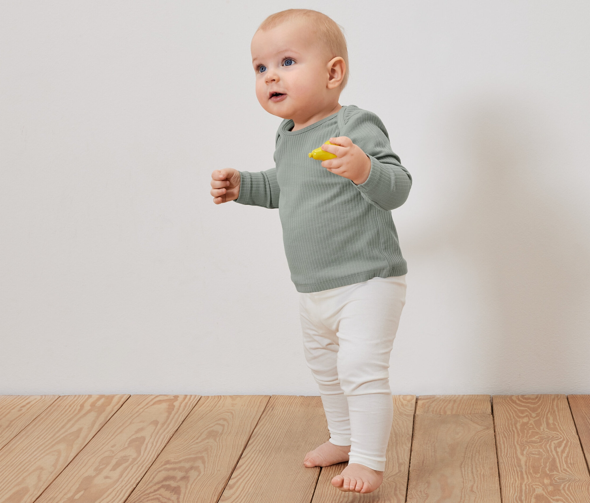 Un bébé debout sur un plancher en bois porte un t-shirt à manches longues côtelé vert clair et un legging beige.
