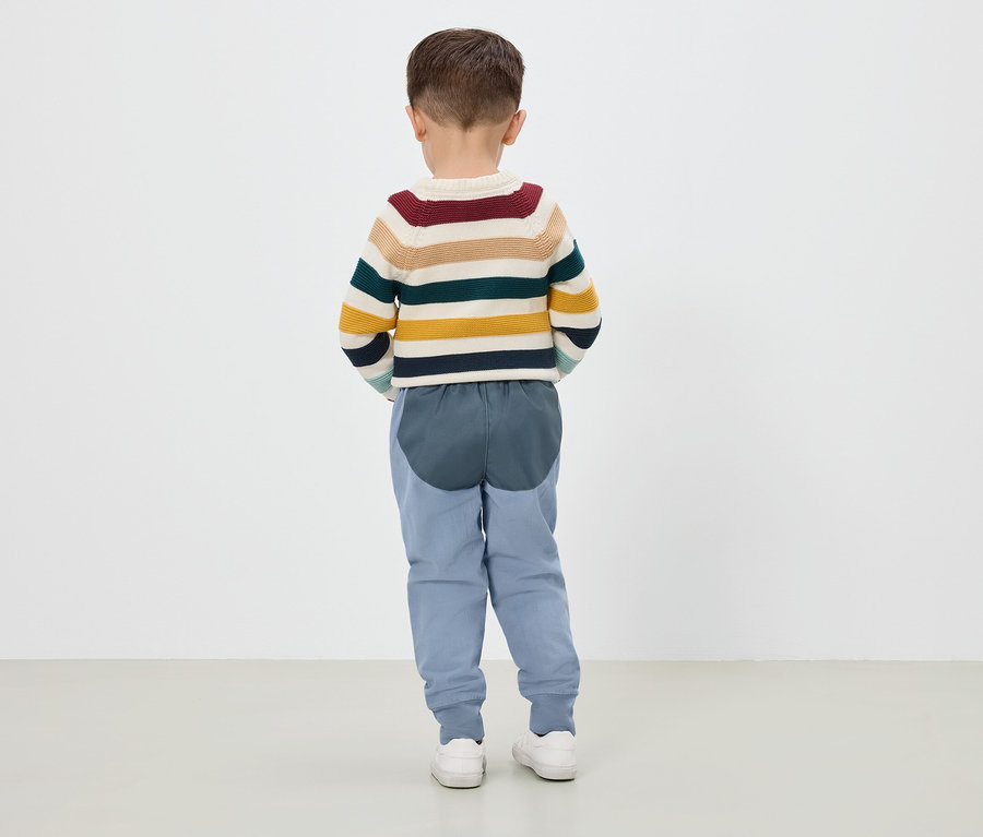 Un garçon debout, vu de dos, porte un pull rayé, un pantalon bleu et des baskets blanches.