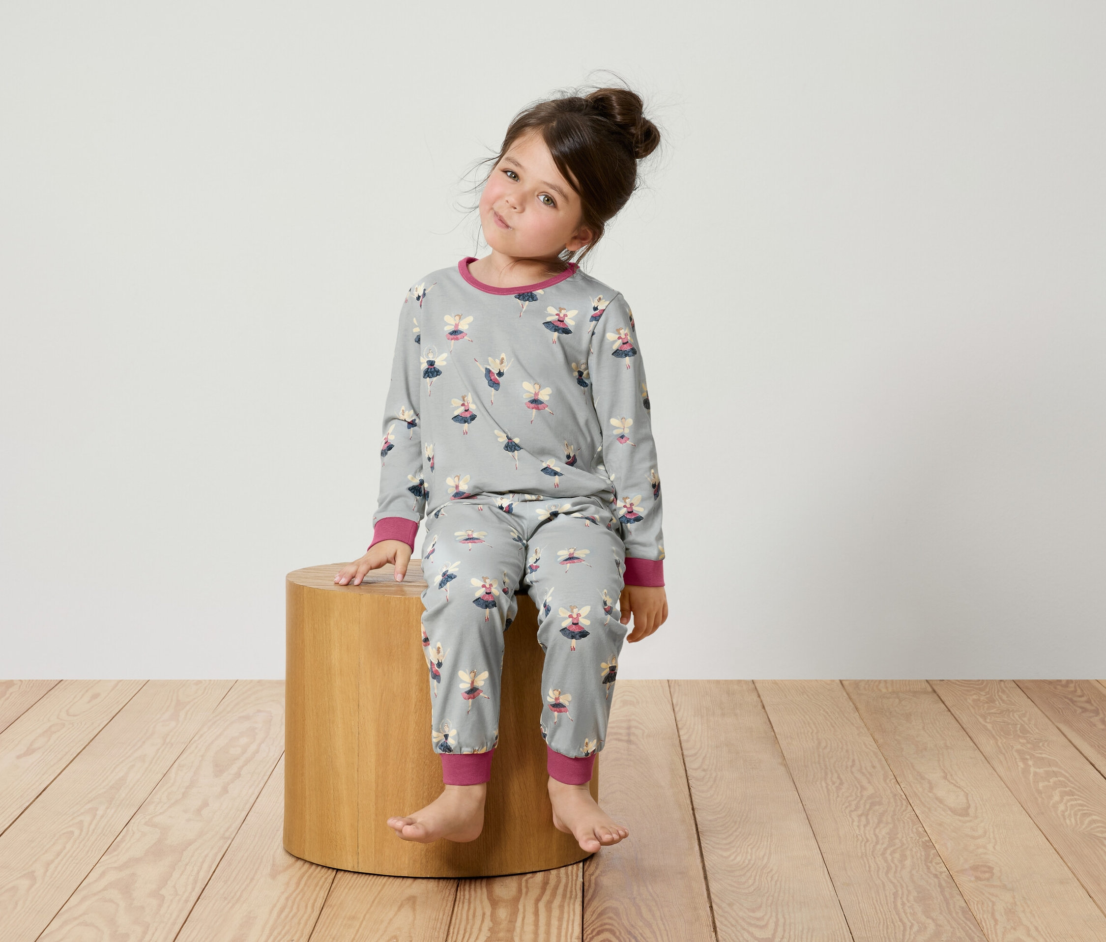 Une fillette est assise sur un tabouret en bois et porte un pyjama gris avec des ballerines.