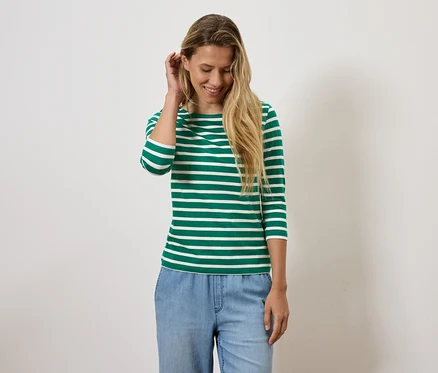 Une femme blonde sourit et ajuste ses cheveux, elle porte un t-shirt rayé vert et blanc et un jean bleu.
