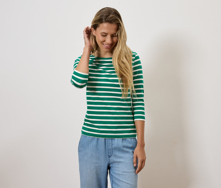 Une femme blonde sourit et ajuste ses cheveux, elle porte un t-shirt rayé vert et blanc et un jean bleu.
