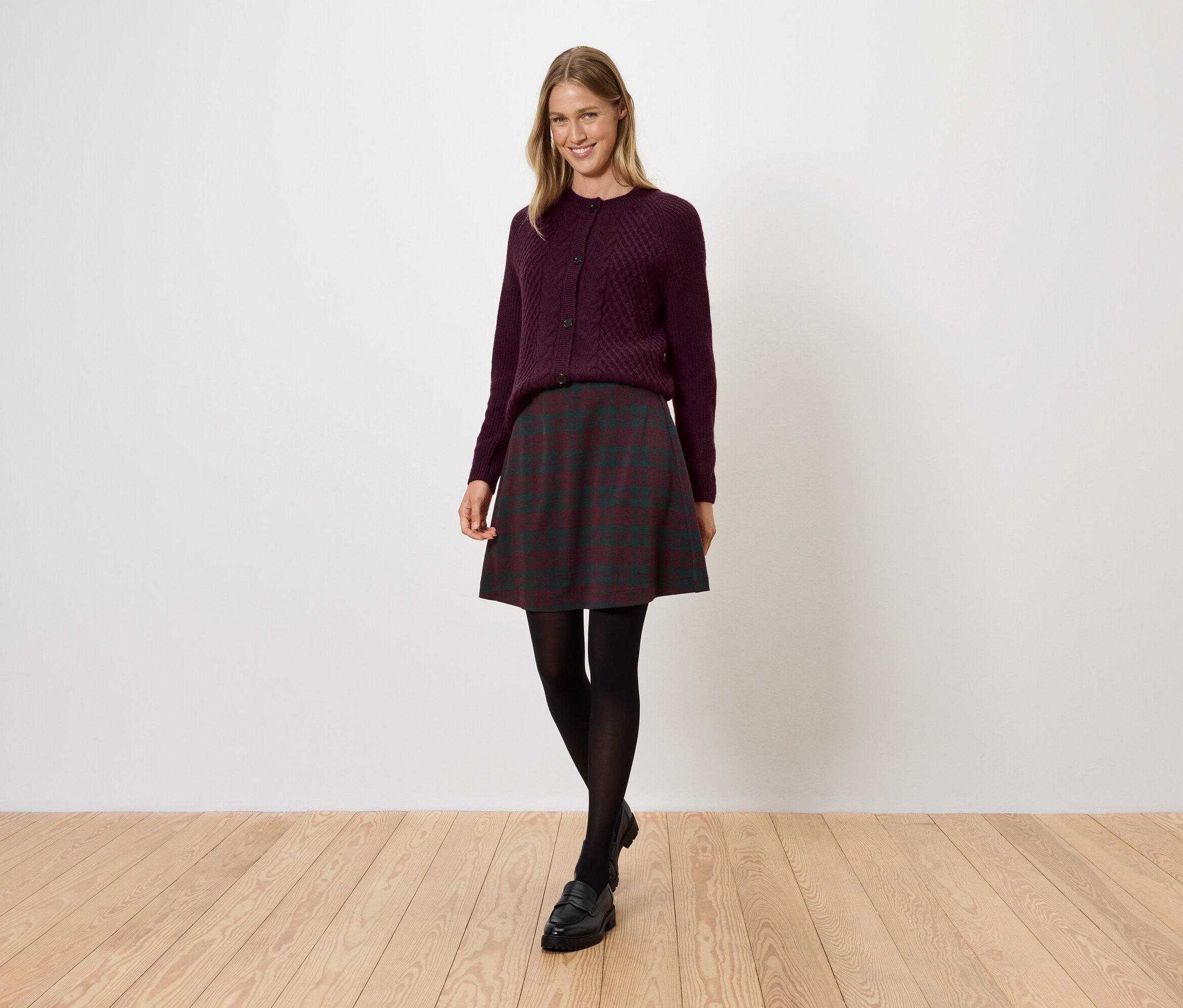 Une femme portant un pull bordeaux, une jupe foncée, des collants noirs et des chaussures se tient sur un plancher en bois.