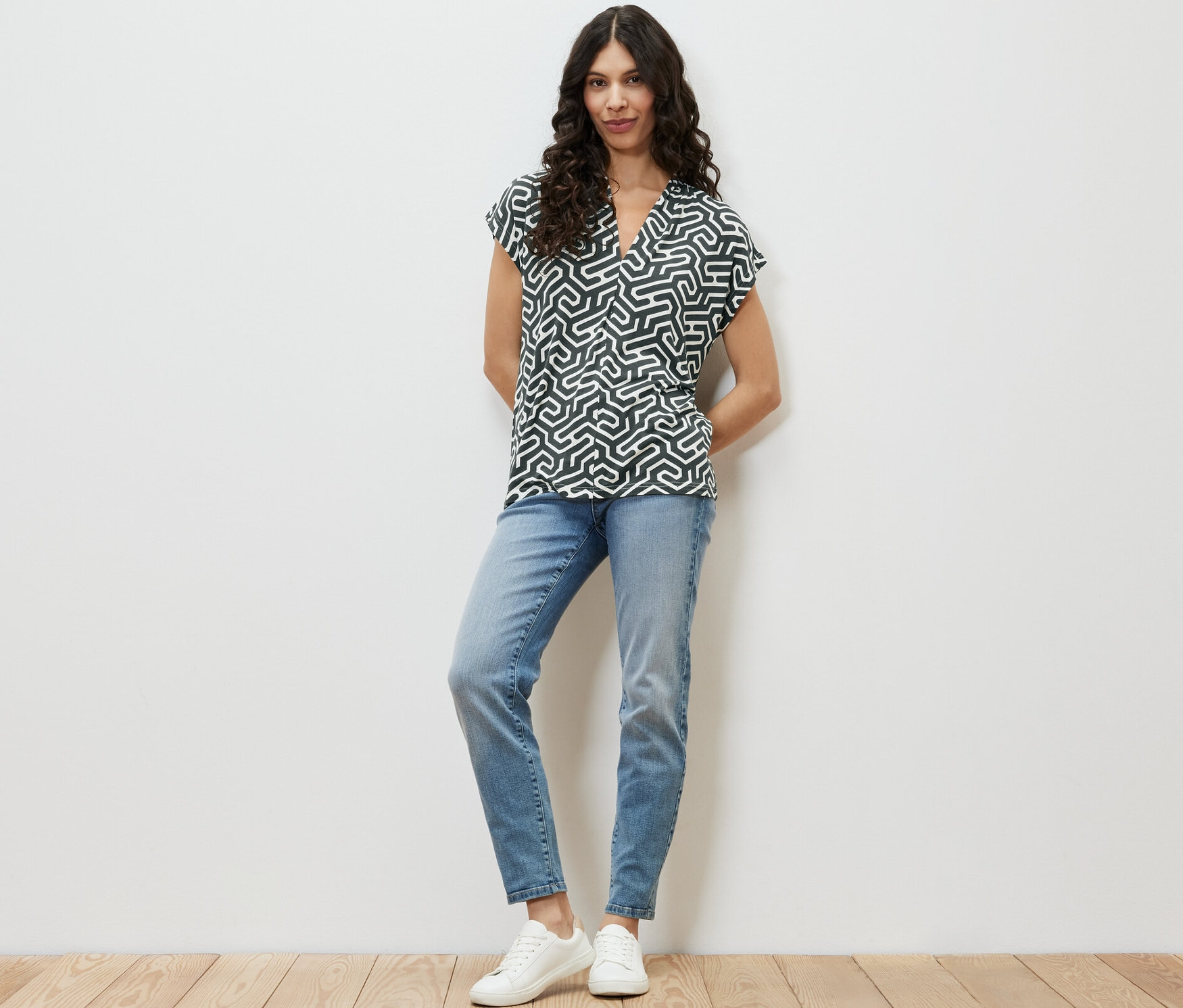 Une femme debout portant une blouse verte à encolure en V, un jean bleu et des baskets blanches.
