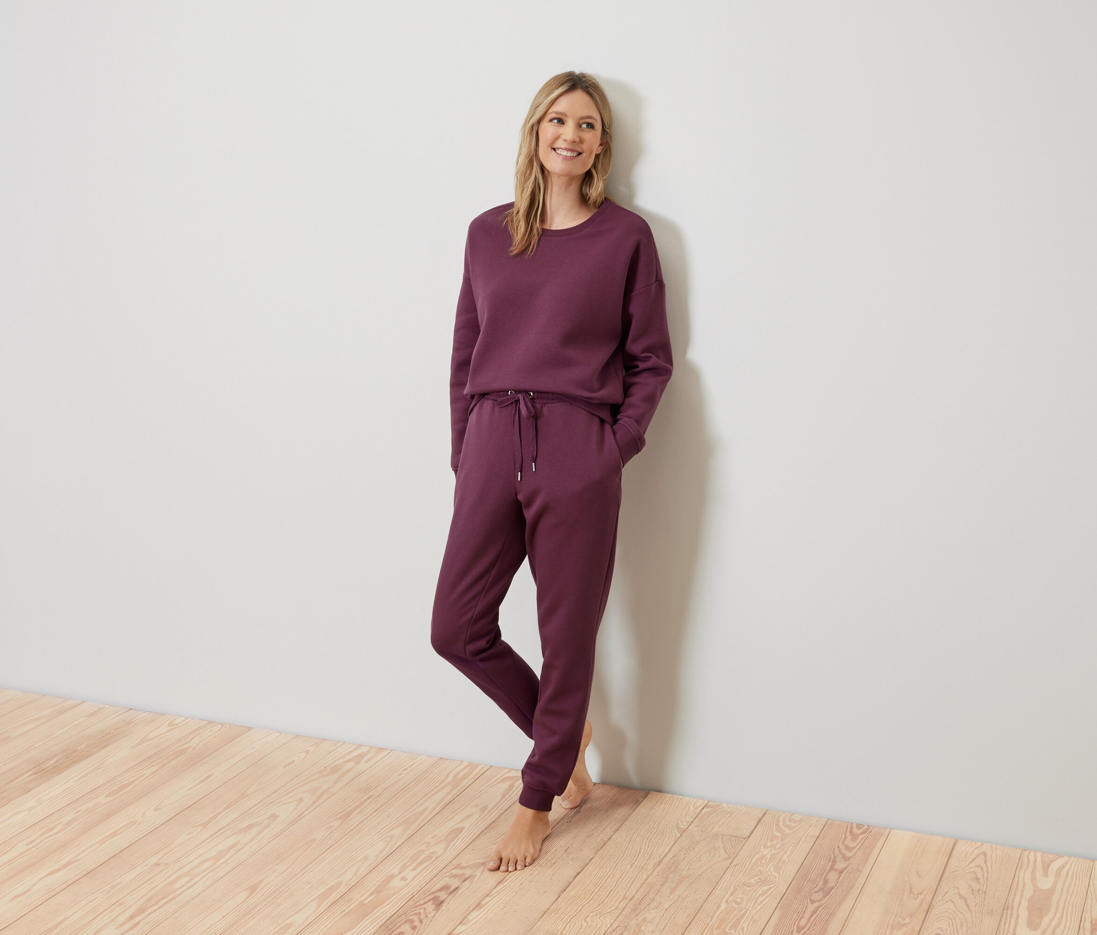 Une femme blonde pose en survêtement bordeaux contre un mur.