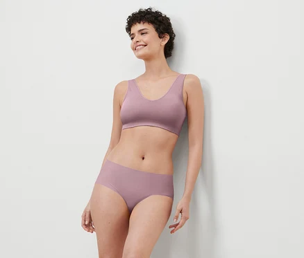 Une femme souriante aux cheveux courts porte un soutien-gorge et une culotte violets et s'appuie contre un mur blanc.