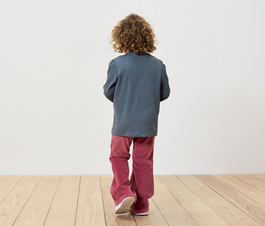 Un garçon aux cheveux bouclés, vu de dos, porte un T-shirt à manches longues bleu-gris et un pantalon bordeaux.