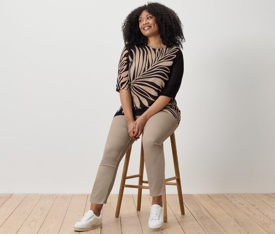 Une femme est assise sur un tabouret et porte un tregging 7/8, un haut imprimé et des baskets.
