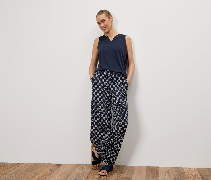 Femme appuyée contre un mur, portant un débardeur bleu marine, un pantalon à motifs et des sandales bleues.