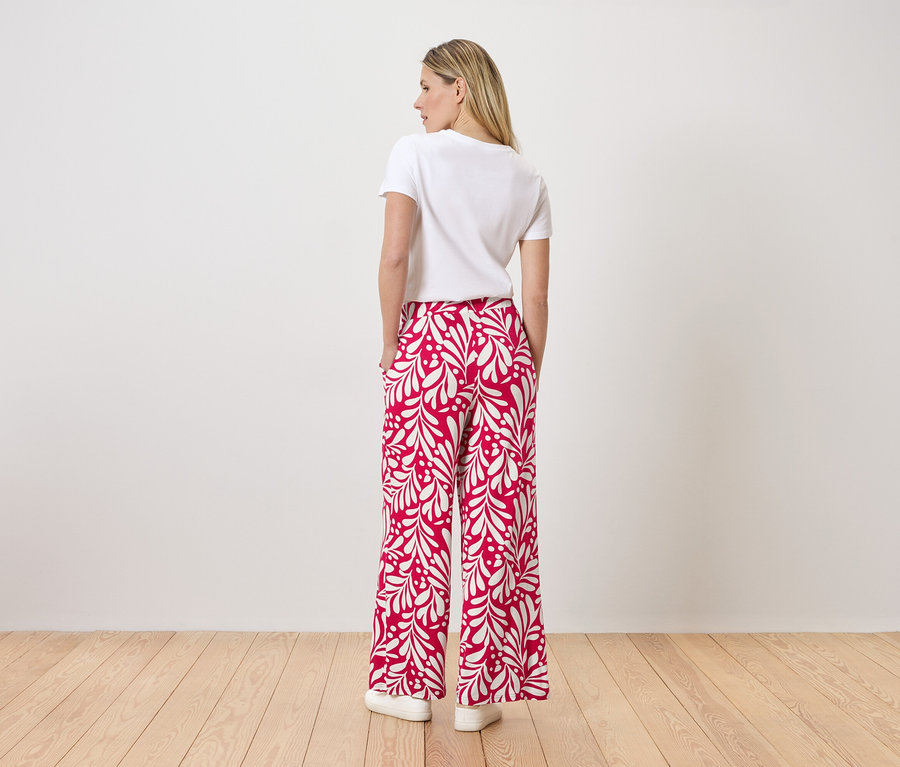 Une femme se tient dos à la caméra, portant un t-shirt blanc et un pantalon rouge avec un motif de feuilles blanches.