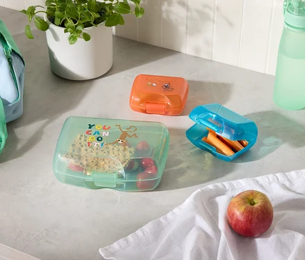 Boîtes à lunch orange, bleues et vertes contenant divers aliments, une bouteille d'eau et un sac à lunch sur un comptoir de cuisine.