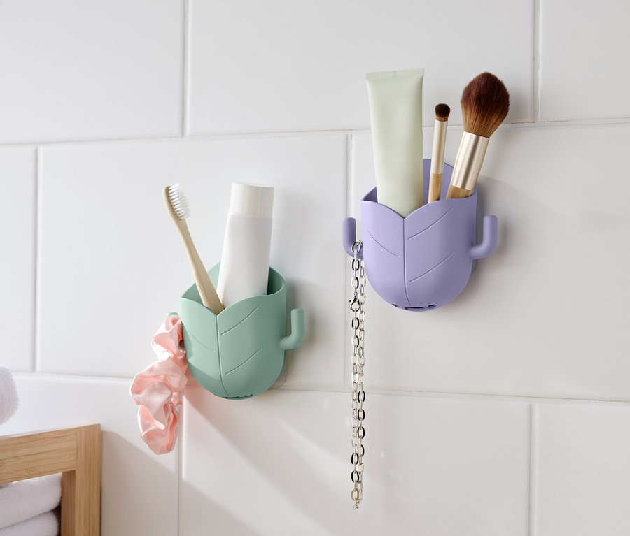Deux récipients en forme de feuille sont accrochés à un mur carrelé blanc. Le récipient de gauche est vert menthe et contient une brosse à dents et un tube. Un chouchou rose est accroché à côté. Le récipient de droite est lavande et contient un tube, un pinceau et une chaîne.
