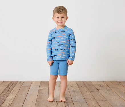 Un jeune garçon se tient pieds nus sur un plancher en bois, portant un t-shirt bleu à manches longues avec des motifs de requins et un short bleu.