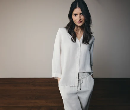 Une femme aux longs cheveux foncés portant un ensemble de pyjama deux pièces clair composé d'une chemise et d'un pantalon, les mains dans les poches.