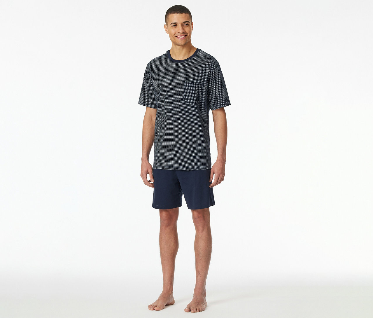 Un homme se tient pieds nus sur fond blanc et porte un lot de pyjama court SCHIESSER bleu marine composé d’un t-shirt à pois à manches courtes et d’un short.