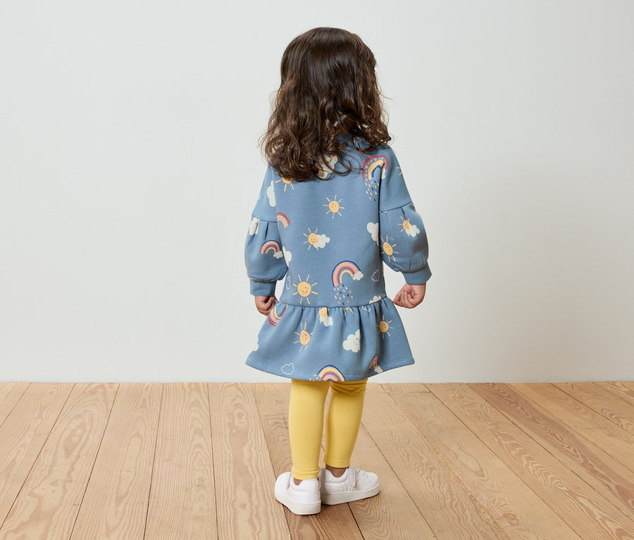 Une petite fille, vue de dos, porte une robe bleue à motifs de soleil, de nuages et d'arcs-en-ciel, des leggings jaunes et des chaussures blanches.
