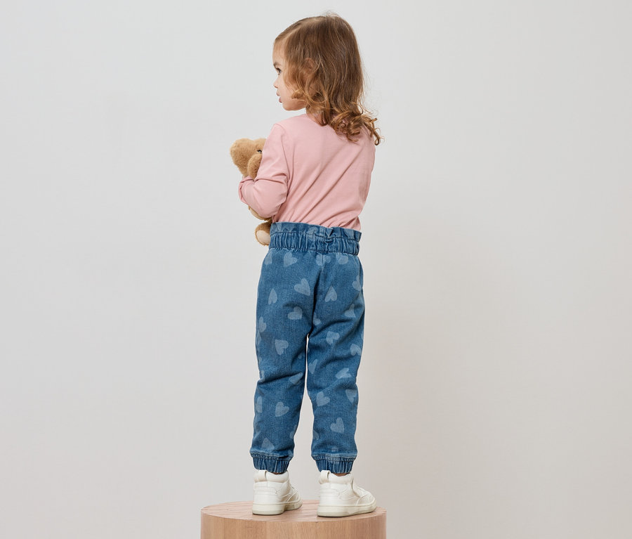 Une fillette aux cheveux bouclés se tient dos à la caméra et serre un ours en peluche. Elle porte un t-shirt rose à manches longues, un jean bleu avec des motifs de cœurs et des baskets blanches.