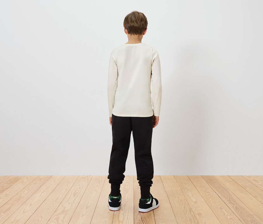 Un enfant se tient dos au spectateur, portant un t-shirt beige à manches longues, un pantalon noir et des baskets vertes et noires.
