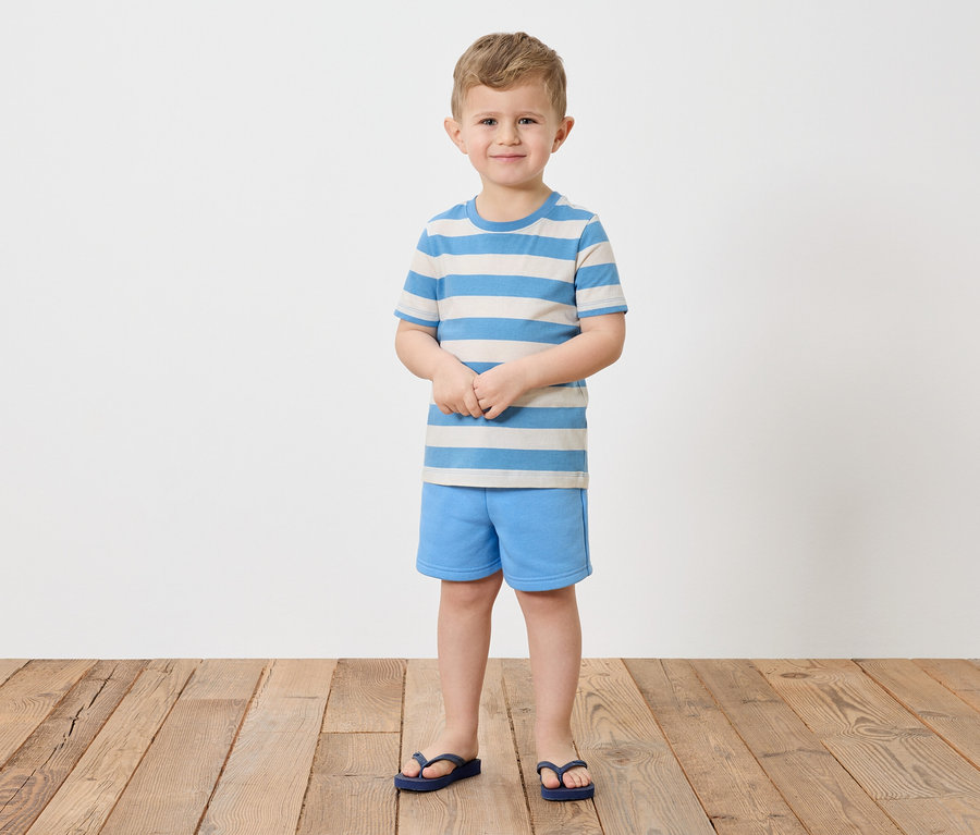 Un garçon aux cheveux blonds se tient pieds nus sur un plancher en bois et porte un t-shirt rayé bleu et blanc, un short bleu et des tongs bleues.