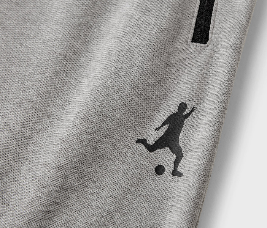 Gros plan d'un pantalon de survêtement gris avec une silhouette noire d'un joueur de football et une poche zippée.