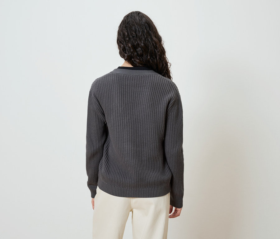 Vue de dos d'une femme aux longs cheveux bouclés et foncés, portant un pull en tricot gris et un pantalon couleur crème.