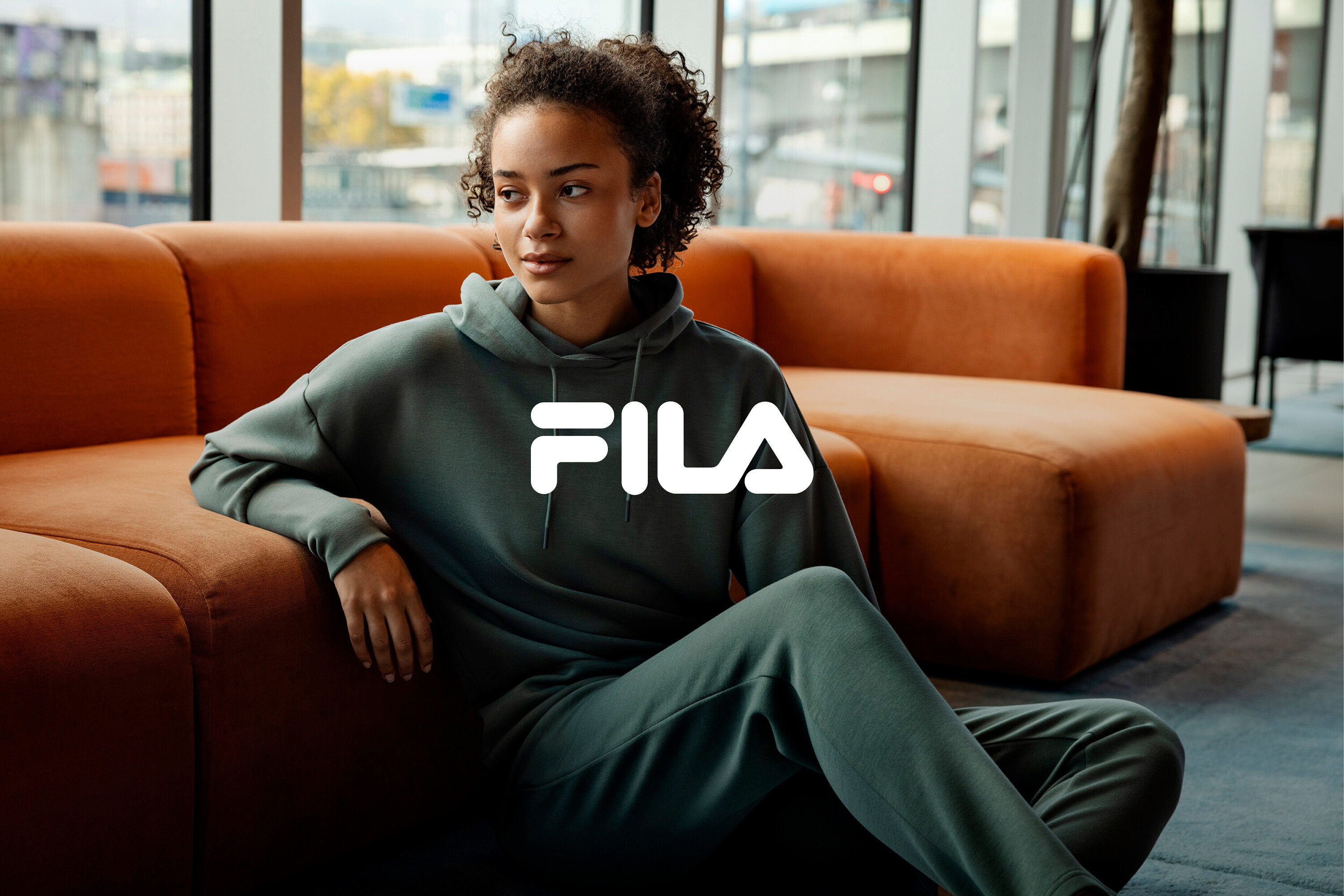 Une femme est assise sur le sol, appuyée contre un canapé orange, portant un survêtement vert Fila.