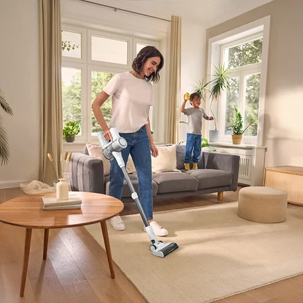 Une femme passe l'aspirateur sur un tapis avec un aspirateur sans fil, un enfant se tient sur un canapé. Table ronde à gauche, fenêtre en arrière-plan.