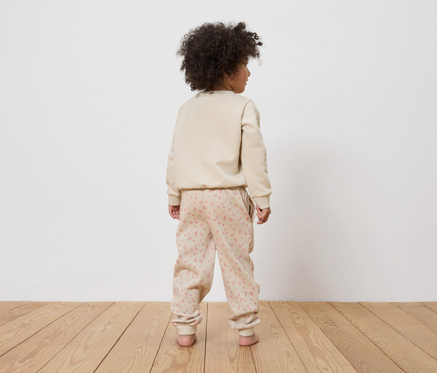 Un enfant aux cheveux bouclés se tient dos à la caméra, portant un pull beige et un pantalon à motifs floraux roses.