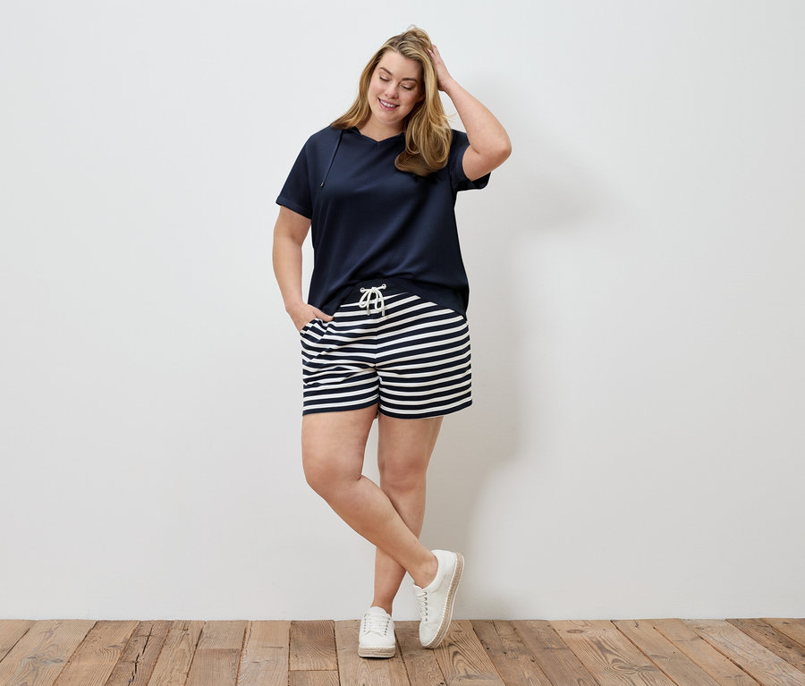 Une femme en t-shirt bleu marine et short rayé pose devant un mur. Elle a une main dans ses cheveux et une dans sa poche.