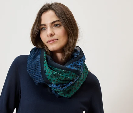 Une femme aux cheveux bruns porte un foulard à motifs bleu et vert sur un fond neutre.