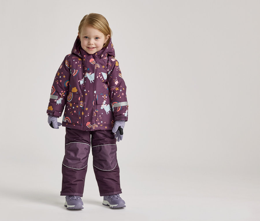 Une fillette se tient sur un fond clair dans une combinaison de ski violette à motifs de licornes et d'arcs-en-ciel, des gants et des chaussures gris.