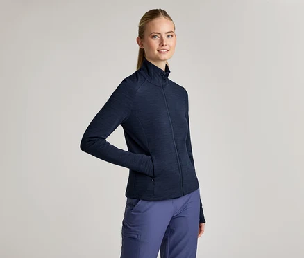 Une femme avec une queue de cheval porte une veste zippée bleu marine et un pantalon bleu marine. Une main est dans la poche de la veste.