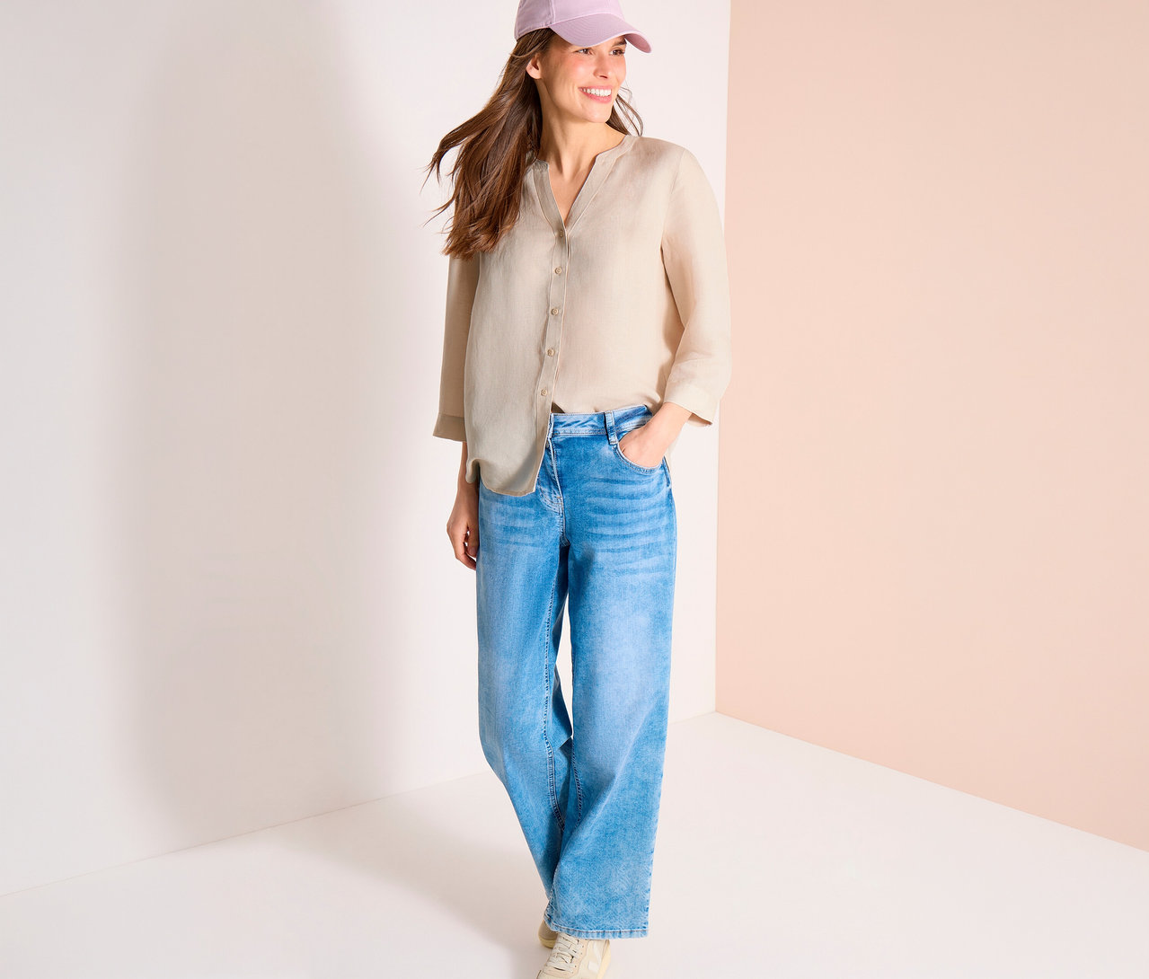 Femme souriante avec une casquette rose, un chemisier beige et un jean bleu avec une main dans la poche.