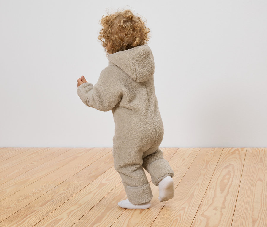 Un enfant aux cheveux bouclés porte une combinaison de bébé en teddy et des chaussettes blanches, debout sur un plancher en bois devant un mur blanc.
