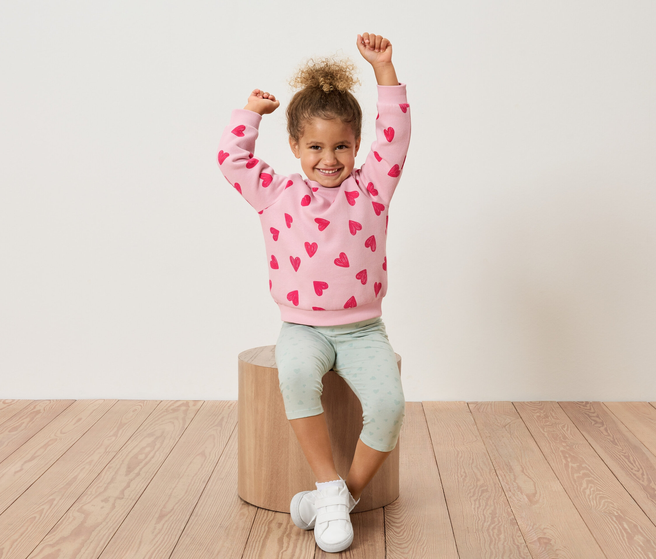 Une jeune fille est assise sur un tabouret en bois, les bras levés, portant un sweat-shirt rose avec des cœurs rouges et un legging vert clair.
