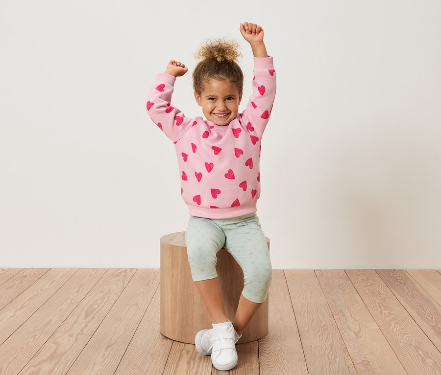 Une jeune fille est assise sur un tabouret en bois, les bras levés, portant un sweat-shirt rose avec des cœurs rouges et un legging vert clair.
