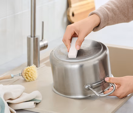 Une personne nettoie une casserole en acier inoxydable avec une éponge abrasive dans un évier, à côté d'une brosse et d'un torchon.