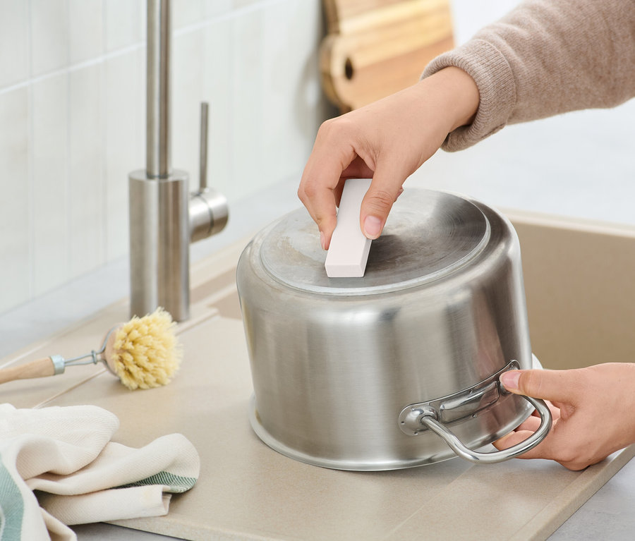 Une personne nettoie une casserole en acier inoxydable avec une éponge abrasive dans un évier, à côté d'une brosse et d'un torchon.